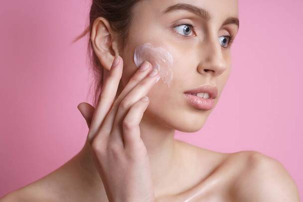 Beautiful young woman applying cream on her face against color background - Zdjęcie, obraz