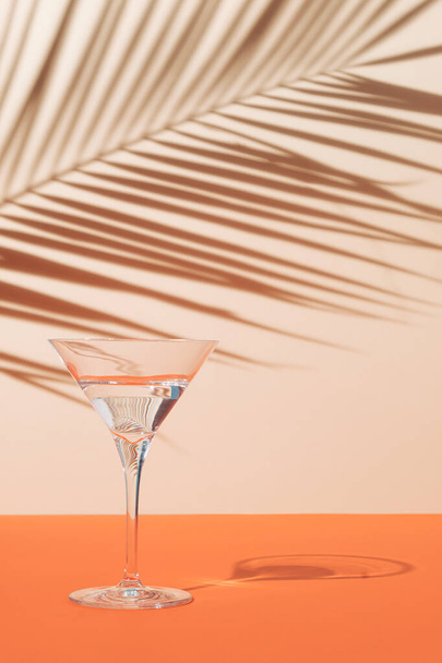 Bovenaanzicht van groen tropisch blad schaduw en martini glas op oranje en zand kleur achtergrond. Zomer tropisch drankje minimaal concept. Elegante samenstelling. - Foto, afbeelding