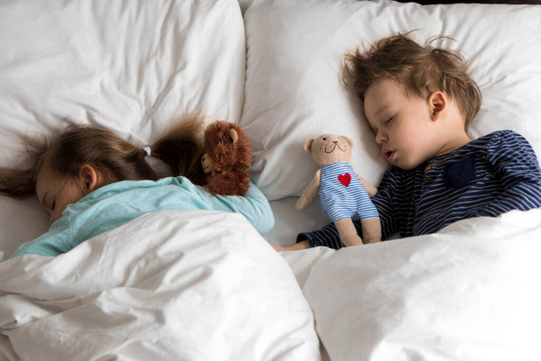 Authentisches Porträt niedlichen kaukasischen kleinen Vorschulgeschwistern Baby Junge und Mädchen im blauen Schlaf mit Teddybär auf weißem Bett. Mittags ruhen die Kinder. Pflege, Medizin und Gesundheit, Kindheit, Lebenskonzept - Foto, Bild