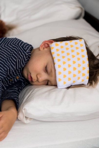 Authentic portrait sick cute caucasian little preschool baby boy in blue sleep with compress on forehead on white bed. child resting at lunchtime. care, medicine and health, Childhood, life concept - Foto, imagen