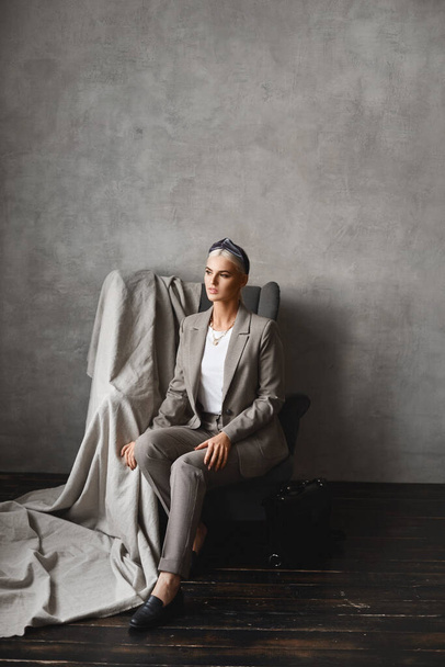 A young Caucasian woman with blond hair looking aside while sitting in the armchair. Gorgeous young businesswoman in stylish suit posing in an empty interior - 写真・画像
