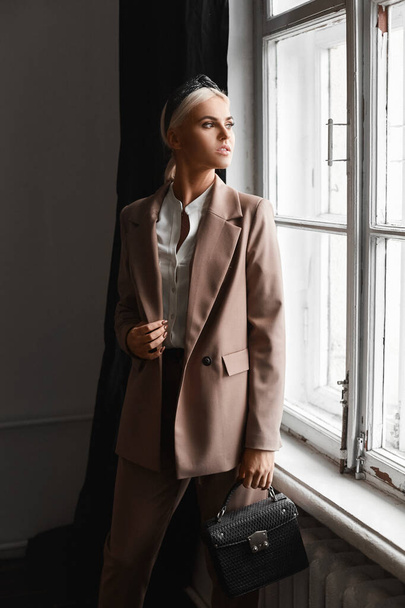 Model girl with blond hair in a modish suit posing with a trendy purse in vintage interior near the window - Photo, Image