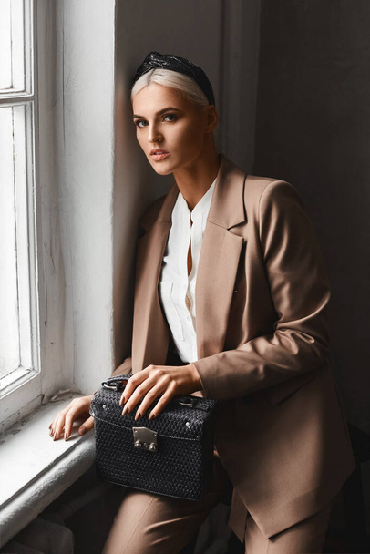 Young model woman with blond hair in a modish suit posing with a trendy purse in vintage interior near the window - Foto, afbeelding