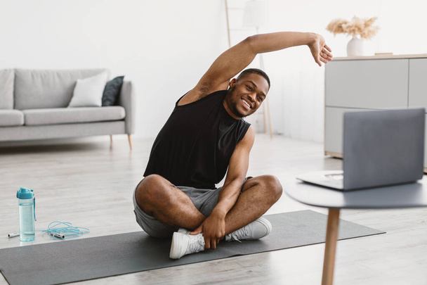 Feliz chico negro haciendo estiramiento en el ordenador portátil haciendo ejercicio en casa - Foto, Imagen