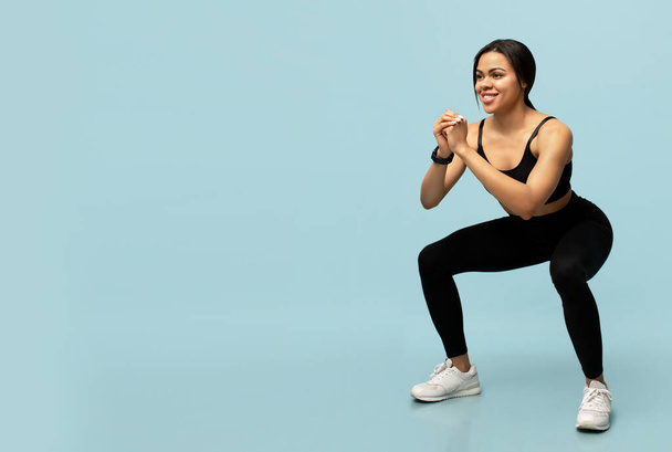 Training concept. Determined black woman doing squats exercise, working out over blue background with empty space - Photo, Image