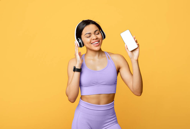 Aplikacja muzyczna do treningu. Podekscytowana młoda czarna kobieta słuchająca muzyki w słuchawkach i trzymająca telefon komórkowy z pustym ekranem - Zdjęcie, obraz