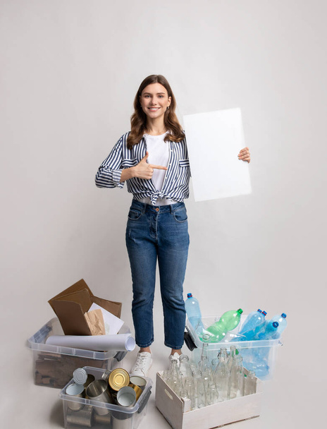Öko-Freiwillige Frau mit leerem Plakat steht zwischen Containern mit unterschiedlich sortiertem Abfall - Foto, Bild