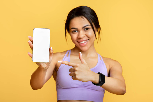 Aplicación deportiva. Emocionada dama negra sosteniendo y mostrando el teléfono inteligente, indicando en pantalla vacía negra para maqueta - Foto, imagen