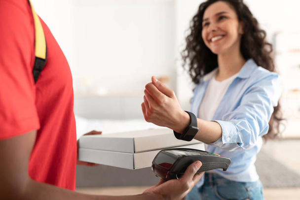 Deliveryman κρατώντας κουτί πίτσα και τερματικό, γυναίκα πληρώνει με SmartWatch - Φωτογραφία, εικόνα