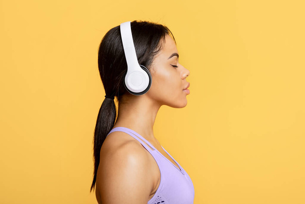 Entrenamiento con música. Mujer afroamericana en uniforme deportivo con auriculares disfrutando de la lista de reproducción con los ojos cerrados - Foto, Imagen