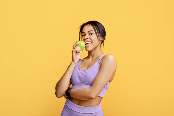 運動後に新鮮な果物を食べ、リンゴをかむよくフィットボディを持つ黒スポーティな女性は、黄色の背景を立って - 写真・画像