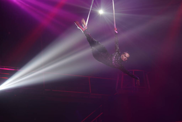 Mujer joven realizando elemento acrobático en correas aéreas contra fondo púrpura oscuro y luz blanca Concepto de club nocturno espectáculo de circo  - Foto, Imagen