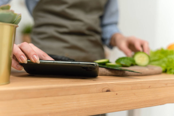 Beautiful woman prepares vegetables in a cozy wooden kitchen looking up a recipe on a smartphone. A young woman browses the website during a cooking lesson. Close-up - Foto, afbeelding