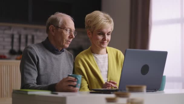 dva důchodci používají notebook doma v kuchyni, surfování na internetu, nakupování on-line, staří muži a ženy zobrazují obrázky na displeji - Záběry, video