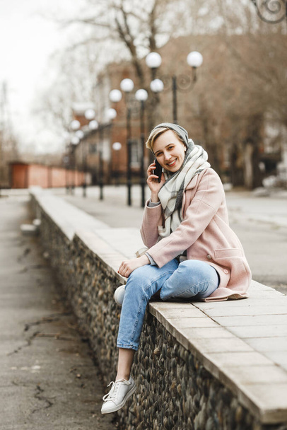 Attractive hipster blonde girl in a pink coat walks around beautiful authentic old town and speaks by phone. The concept of freedom. creativity and inspiration - Φωτογραφία, εικόνα