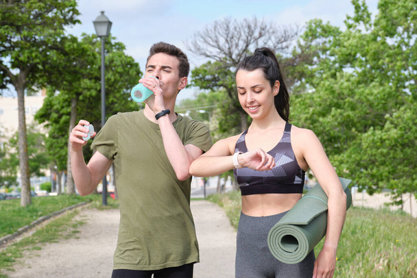 公園に立っている間、フィットネススマートウォッチと飲料水を見ている若いスポーツカップル。フィットネス技術の概念. - 写真・画像