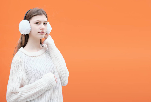 Módní teenager dívka na sobě bílé chrániče sluchu se svetrem a stojí proti oranžové pozadí. - Fotografie, Obrázek