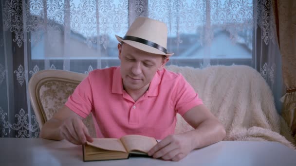 Un homme dans un chapeau le soir lit un livre intéressant assis à une table - Séquence, vidéo