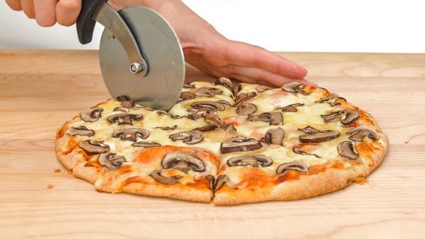 Femme coupe une pizza aux champignons à l'aide d'un coupe-pizza, gros plan sur une planche à découper en bois sur fond blanc - Photo, image