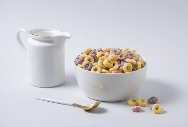 flocons de maïs colorés doux anneaux dans un bol blanc avec cuillère et une cruche de lait sur un fond gris. Vue de face et copier l'image de l'espace - Photo, image