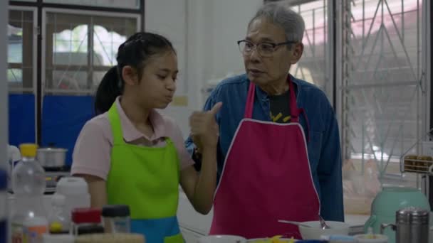 Здоровый азиатский пожилой дедушка в фартуке кормит кислым манго с вкусным соусом, приготовленным для его очаровательной внучки в кухонной комнате. Связывающие отношения в семье.  - Кадры, видео