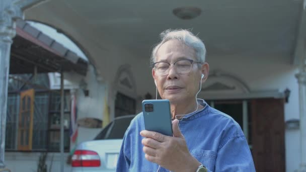 ポートレートアジアの高齢者は、家の前に立っている間にスマートフォンからビデオ通話カメラで友人に伝えられたイヤホンを身に着けて引退した男。現代のテクノロジーライフスタイル. - 映像、動画