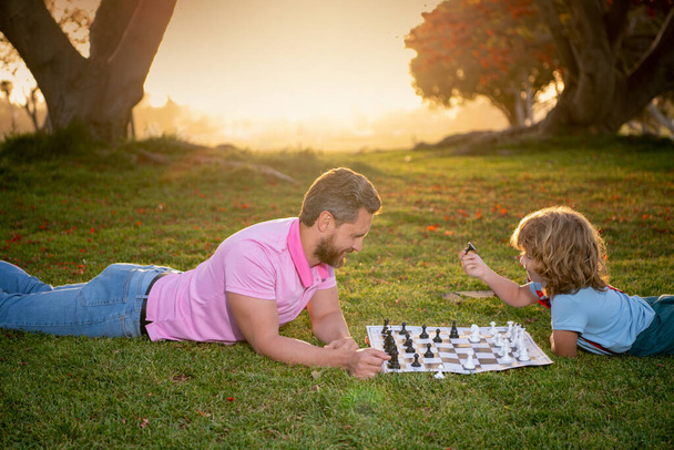Son laying on grass and playing chess with father on sunset in sunny park. - Фото, зображення