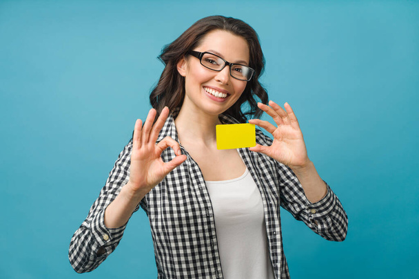 Linda mujer sonriente en camisa de vestir y gafas, mostrando la tarjeta de crédito en la mano para el concepto de la sociedad financiera y sin efectivo, posando sobre fondo azul. Copiar espacio. - Foto, imagen