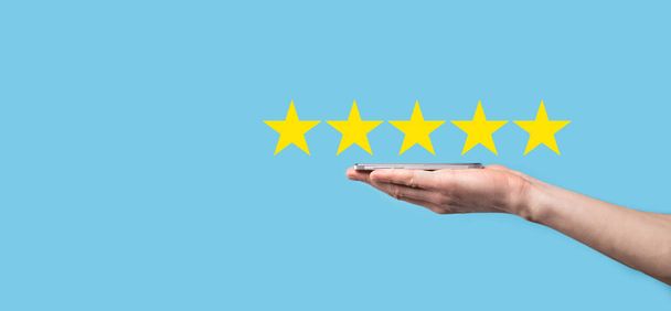 Człowiek trzyma smartfon w rękach i daje pozytywną ocenę, ikona pięć symbol gwiazdy, aby zwiększyć rating koncepcji firmy na niebieskim tle.Customer service experience and business satisfaction survey - Zdjęcie, obraz