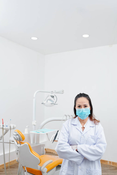 Kendine güveni tam, koruyucu maskeli dişçi portresi. Dişçi koltuğunda duruyor. - Fotoğraf, Görsel