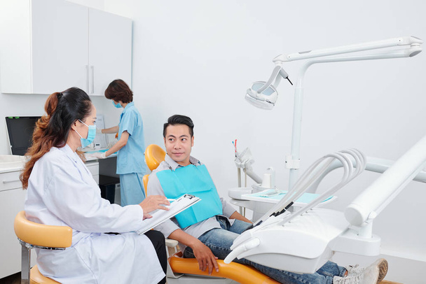 Odontóloga hablando con paciente y llenando documento médico cuando la enfermera prepara herramientas en segundo plano - Foto, imagen