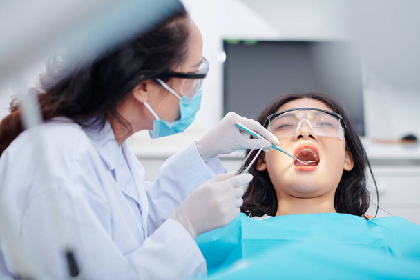 Οδοντίατρος που χρησιμοποιεί οδοντικό καθρέφτη και τσιμπιδάκι κατά τον έλεγχο των δοντιών της νεαρής γυναίκας ασθενή - Φωτογραφία, εικόνα