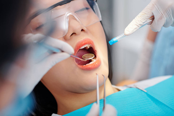 Zbliżenie obrazu dentysty z lustrem i wyrzutnikiem śliny badającym zęby młodej pacjentki - Zdjęcie, obraz