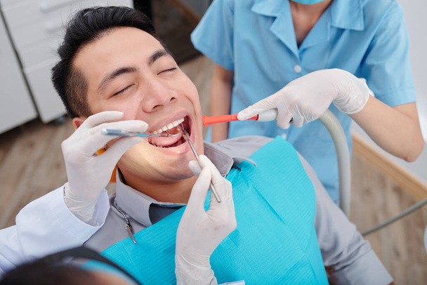 Дантист лечит зубы пациента-мужчины, когда медсестра кладет эжектор слюны внутрь рта - Фото, изображение