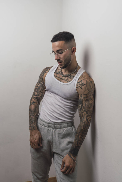 A vertical shot of a good looking tattooed young European man leaning on a white wall - 写真・画像