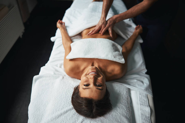 Positive joyful lady feeling great while unrecognized male masseur stroking client body in spa salon - Фото, изображение