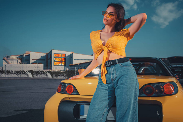Kobieta w niebieskich dżinsach, pomarańczowym top i okulary przeciwsłoneczne jest pozowanie w pobliżu żółty samochód kabriolet z papierową filiżanką kawy na bagażniku na parkingu. Przestrzeń kopiowania - Zdjęcie, obraz