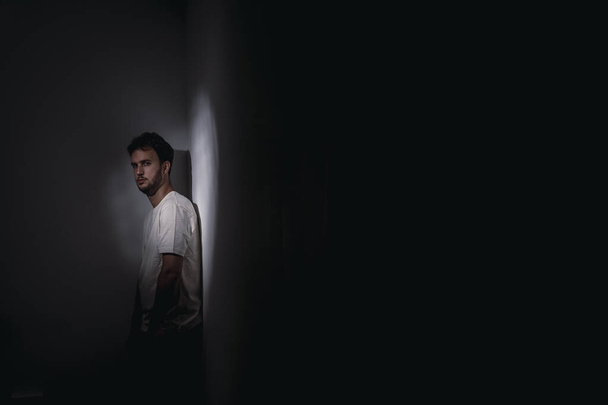 A young Spanish male in a white t-shirt standing at the corner of a dark room leaning on the wall under dim lighting - Fotó, kép