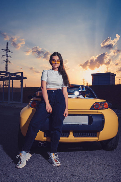 Žena ve slunečních brýlích, bílém svršku, modrých džínách a teniskách pózuje venku u žlutého auta se dvěma skleněnými lahvemi limonády na kufru. Vysmát - Fotografie, Obrázek