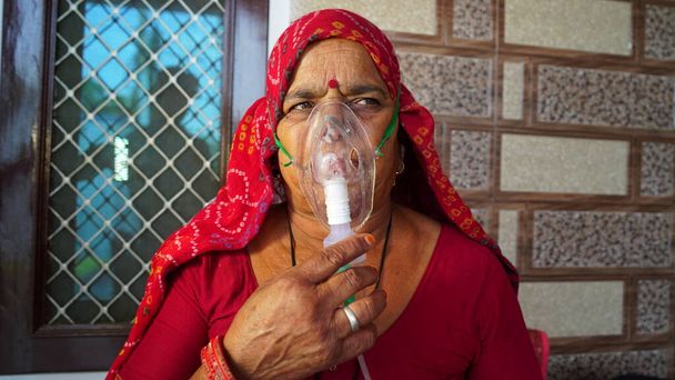 Old Indian woman infected with Covid 19 disease. Patient inhaling oxygen wearing mask with liquid Oxygen flow. - Photo, Image