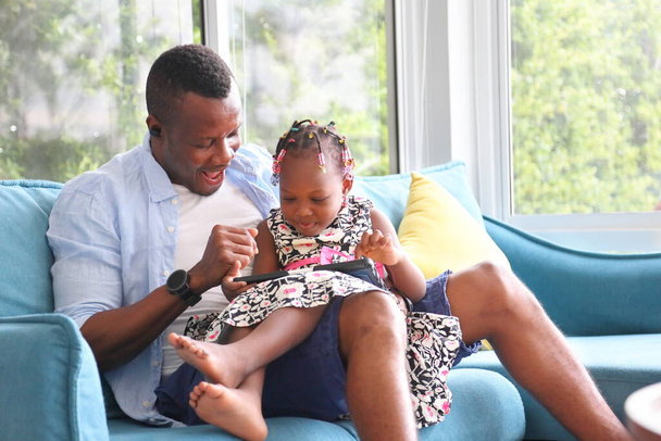 Афроамериканець шукає свою маленьку дочку, яка вчиться на цифровому планшеті з дому на дивані. - Фото, зображення