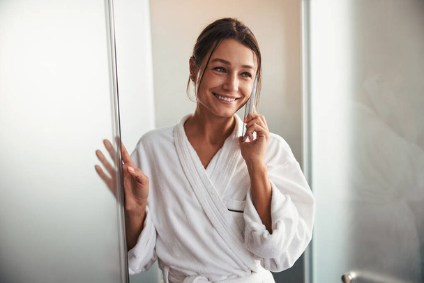 Brunette Caucasian charming business woman in white bathrobe having dialogue on cell phone in beauty salon - Fotó, kép