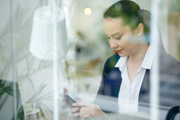 Молодая улыбающаяся деловая женщина проверяет текстовые сообщения в смартфоне, сидя за журнальным столиком, просматривая через окно - Фото, изображение