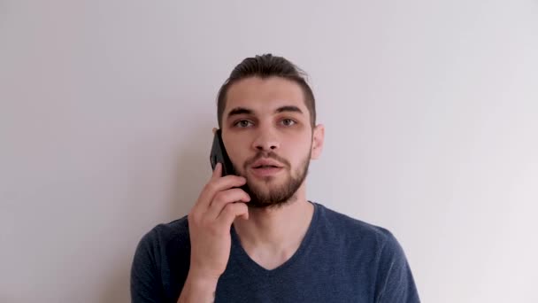 Young Caucasian man with beard and stylish hairstyle in blue Tshirt on white background is talking on phone, angry and irritated. Unpleasant conversation on phone. - Footage, Video