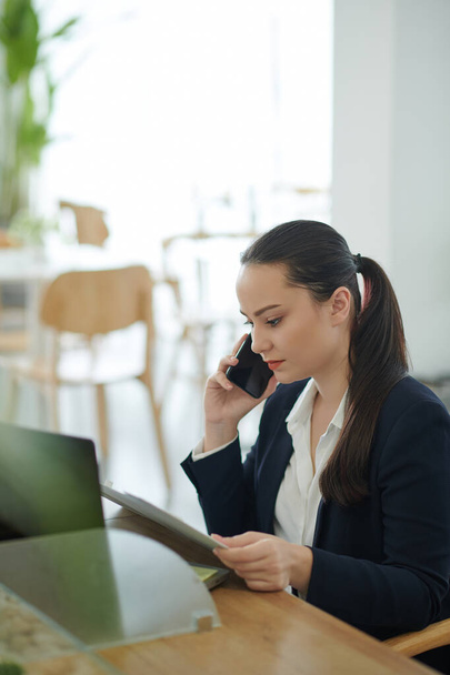Portret poważnej młodej kobiety przedsiębiorcy rozmawiającej przez telefon i czytającej dokument o rozwoju biznesu - Zdjęcie, obraz