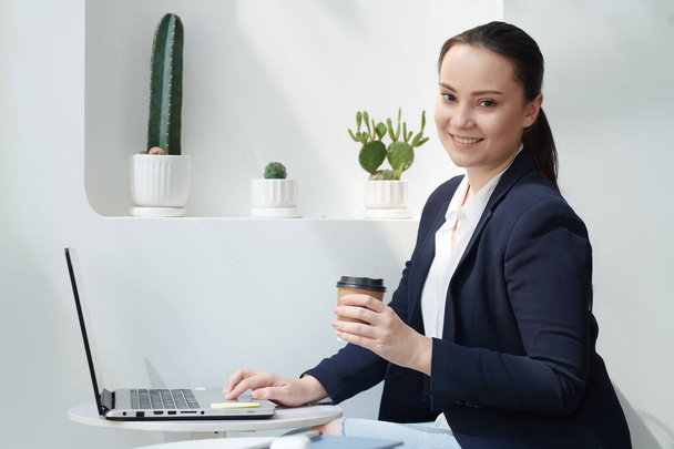 Porträt einer lächelnden jungen Geschäftsfrau mit einer Tasse Kaffee zum Mitnehmen, die am Cafétisch sitzt und am Laptop arbeitet - Foto, Bild