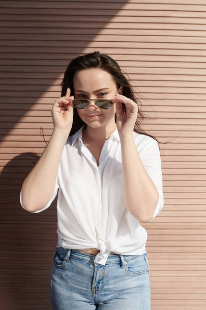 Beautiful smiling brunette in white shirt taking off sunglasses and looking at camera - Fotoğraf, Görsel