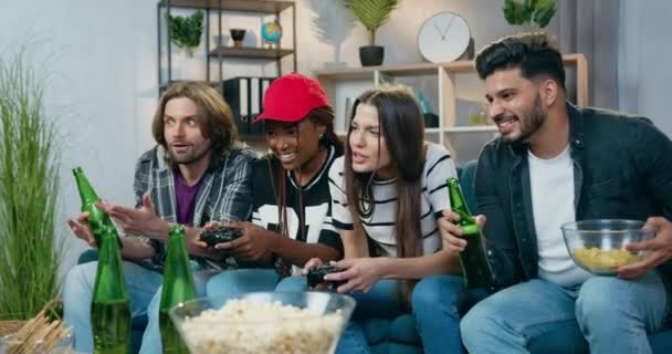 Handsome emotional two guys sitting on the couch together with mixed race girls which competing in videogames on TV set applying gamepads ,leisure concept - Filmmaterial, Video