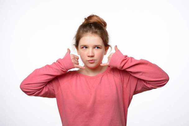 Call me later. Pensive young girl makes phone gesture, pretends mobile communication, wears casual pink sweatshirt. Body language concept. Studio shot, white background - Photo, image