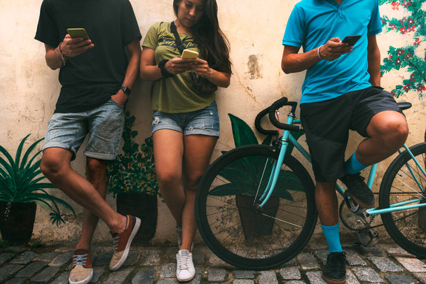 παρέα φίλων στο δρόμο με ένα ποδήλατο απασχολημένο ο καθένας με το κινητό του τηλέφωνο στοιβαγμένο σε έναν τοίχο βαμμένο κίτρινο με φυτά. ομάδα νέων ανθρώπων που ασχολούνται ο καθένας με το smartphone του. φόντο με copyspace. έννοια τρόπου ζωής. έννοια της τεχνολογίας. - Φωτογραφία, εικόνα